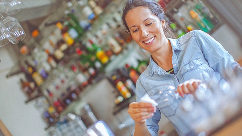 A woman standing behind a bar cleaning a wine class with a cloth